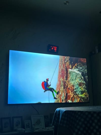 An indoor residential scene in Topolchane, Bulgaria, is depicted under dim lighting, suggesting evening hours. A large, dark-framed television screen dominates the center, displaying an image of a person rappelling down a rocky cliff face. The individual wears a helmet with a mounted camera or headlamp, goggles, a blue jacket, a red backpack, green leggings, and black shoes, actively descending a rope against a bright blue sky on the left and a multi-hued rock formation on the right. Above the television, a red LED digital clock is mounted, displaying the date "1 2 2020" and the time "21:19:21". Below the television, a shelf holds several framed pictures and various small objects. To the right, a corner of a table covered with a dark, light-checkered tablecloth is visible. The TV screen is the primary light source within the room.