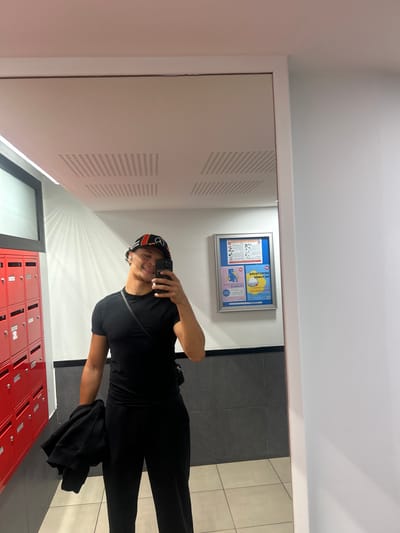 A person is captured in a mirror selfie within an indoor hallway or lobby, located in Noisy-le-Grand, France. The individual, appearing male, is seen from the waist up, holding a black smartphone to take the photo. They are smiling broadly at the camera. The person is wearing a black t-shirt, black trousers, and a black, orange, and white cap. A black jacket or sweatshirt is casually held in their left hand.
The setting is a well-lit space with white walls and ceiling. To the left of the person (in the reflection), there are perforated panels or ventilation grates on the wall. Directly below the person, a bank of red mailboxes or lockers with silver locks is visible. In the background, behind the person's reflection, there is a framed blue bulletin board displaying various notices and diagrams, including text that appears reversed due to the mirror, with fragments like "DE L'ANNEE" and "JANDI KUDUA" partially legible. The floor is tiled, featuring dark grey tiles on one side and lighter, possibly beige, tiles on the other. The overall impression is a modern, communal area within a building.