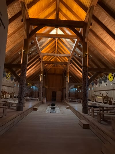 This media file depicts the interior of a large, traditional wooden building, likely a longhouse, with a steeply pitched, timber-framed roof. The structure is constructed entirely of wood, with large, exposed beams forming the ceiling and supporting the roof. The overall aesthetic suggests a historical or replica Viking-era setting.
The scene is characterized by its warm, wooden tones, illuminated by artificial lighting that creates a cozy and somewhat dim ambiance. The camera angle is looking upwards and down a long hall.
Notable objects include several decorative, circular shields with colorful patterns hanging from the ceiling at intervals along the hall. Two large, ornately carved wooden pillars are prominently featured, their surfaces covered in intricate, swirling designs. In the distance, down the length of the hall, more of these decorative pillars and shields are visible, along with what appear to be wooden benches or seating areas and other wooden furniture. A barrel is visible on a raised platform to the right.
The image does not show any people, nor is there any visible text. The time of day is not discernible, and there is no indication of weather conditions as the scene is entirely indoors. The location context of Allerslev, Denmark, combined with the architectural style and decorative elements, strongly suggests a historical reenactment site, museum exhibit, or a building inspired by Viking architecture. The absence of people and activity gives the scene a quiet, almost contemplative feel, focusing entirely on the impressive craftsmanship and historical atmosphere of the architecture.