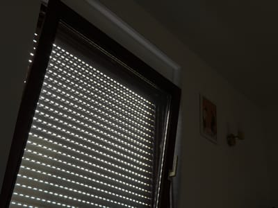 The image shows a closed window with horizontal blinds partially covering it, allowing thin beams of light to pass through. The window frame is dark brown. To the right of the window, on a cream-colored wall, hangs a framed picture depicting two figures. Below the picture, a light fixture is mounted on the wall. The scene is dimly lit, with shadows obscuring much of the room. The image was taken in Kirtorf, Germany.