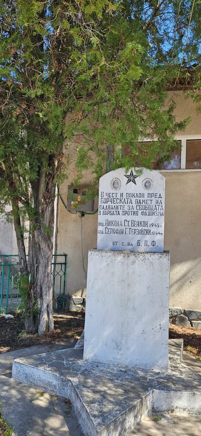 The image shows a memorial monument in Slatino, Bulgaria, dedicated to those who fought against fascism. The monument has a grey top section with an inscription in Bulgarian, two portrait icons, and a star symbol. The inscription honors the memory of fighters who fell for freedom. The names "ПОД. НИКОЛА СТ. ВЕНКОВ" and "ПОД. СЕРАФИН Г. ТЕРЗИЙСКИ," with the years 1945 and 1944, respectively, are inscribed, suggesting they were individuals commemorated by the monument. The monument is set against a building with a window and an electrical box is mounted to the building. A tree stands nearby, obscuring parts of the monument. A metal fence borders the base of the monument. The monument is on a concrete foundation, and there is a sidewalk nearby.