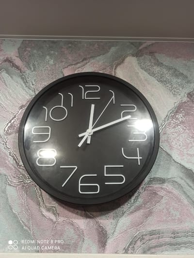 A black analog clock is mounted on a textured wallpaper with pink and grey marbled patterns. The clock face is black with white numbers and white hands. The hour hand is pointing at the 12, and the minute hand is pointing just past the 12, indicating the time is approximately 12:02. The image is taken from a slightly elevated angle, looking down at the clock. The bottom left corner of the image contains a watermark that reads "REDMI NOTE 8 PRO AI QUAD CAMERA". There are no people or any other objects visible in the frame. The location of the image is Kugesi, Russia.
