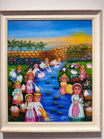 The image is a painting depicting a scene with several young girls in traditional clothing by a river. The girls are engaged in various activities, some are washing clothes in the river, others are carrying baskets, and one is standing in the foreground holding a pink cloth. The background features a brick wall, palm trees, and a sunset sky with orange and yellow hues. The painting style is vibrant and somewhat naive. The signature "A. Violán" is visible in the lower right corner. The painting is framed in a light-colored, textured frame. While the image itself does not contain information about its location in La Orotava, Spain, the style of painting, particularly the traditional attire of the figures and the depicted landscape, is consistent with folk art from the Canary Islands, where La Orotava is located.