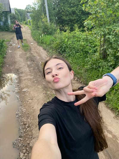A young woman is taking a selfie with a pout and her fingers in a peace sign gesture. She is wearing a black t-shirt and has long brown hair. A blue watch is visible on her wrist. In the background, a dirt road with puddles of water and green vegetation can be seen. Another person, a young man wearing a black t-shirt and shorts, is walking away in the distance on the road. The scene appears to be outdoors, possibly in a rural or suburban area. The lighting suggests it might be daytime, perhaps late afternoon given the shadows. There is no visible text in the image.
