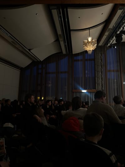 The image shows a darkened room with a large audience seated facing a stage in the rear. The audience is primarily composed of adults, with a few children seated near the front. The room has a modern design, with a curved ceiling and large windows that are covered in curtains. A single chandelier hangs in the center of the room, its light illuminating the surrounding area. There is a spotlight shining from the stage. The scene is lit only by the chandelier, the spotlight, and the ambient light from outside. The room is located in a building on the outskirts of Bingen am Rhein, Germany (49.969, 7.892). The room is part of the Rheingau Musik Festival, a well-known classical music festival. The audience is attentive, and some are taking pictures. The event appears to be a concert or performance.