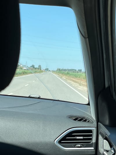 The image is taken from inside a car looking out the windshield. The foreground shows the dashboard with a textured surface and air vents. The windshield reveals a road with white lane markings stretching into the distance under a clear blue sky with faint power lines. On either side of the road, there are grassy areas and vegetation. In the distance, there are some trees and a few small buildings. The overall scene depicts a drive on a sunny day along a highway outside of the city of Ilyinka, Russia.