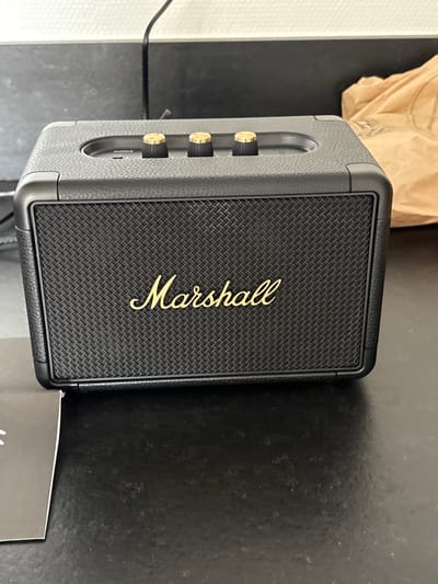 A black Marshall-branded portable Bluetooth speaker is prominently displayed, centered and facing forward, resting on a dark, reflective surface within an indoor setting in Blois, France. The speaker features a textured black "leatherette" exterior, a woven black grille with a prominent gold "Marshall" script logo across its center, and three brass-colored knurled control knobs aligned horizontally on its top surface. A smaller power indicator or switch is visible between the first two knobs. In the blurred background, to the right of the speaker, a crumpled brown paper bag is visible. To the bottom left foreground, a corner of a white instruction manual is partially visible. The background also includes a light-colored wall section with a dark cable running down, and an indistinct dark vertical surface. No individuals are present, and no active interactions are depicted.
