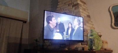 The image shows a television screen displaying a scene from a show. Four people are visible: a man in a dark jacket, a man in a dark coat, a woman in a dark coat, and a man in a dark suit. They are standing in a room and conversing. The television is mounted on a wall with a brick fireplace behind it. There is a plastic bag, a candle holder, and a ceramic vase on the mantle. The scene takes place in a residential home located at the coordinates -36.57, -56.696 in Mar del Tuyú, Argentina. The image is taken from a low angle perspective.
