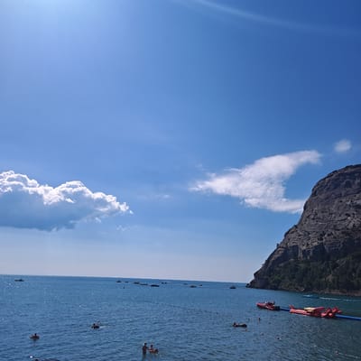 The image depicts a scenic view of the Black Sea near Novyi Svet, Ukraine. A large, rocky cliff face, densely covered with dark green vegetation, dominates the right side of the frame. The cliff meets the calm, blue water at its base. The sea is dotted with several small boats and personal watercraft, including red inflatable structures that appear to be water toys or small boats. A blue barrier or buoy line stretches across a portion of the water. Several individuals are visible in the sea, some swimming, and at least two are in the shallow water at the bottom of the frame, appearing to be interacting. The sky above is a vibrant blue, with scattered white clouds, one large cumulus cloud on the left and a wispy trail of clouds near the center. The sun is bright, casting a slight glare on the water's surface. The overall impression is a sunny day at a coastal recreational area.