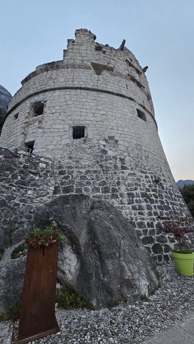 The image captures a weathered, stone-built tower, possibly a historic fortification, set against a pale blue sky with hints of a setting sun visible in the upper right. The tower's circular structure is evident, with rough-hewn stones forming its walls, some of which appear to be in disrepair. Small, barred windows punctuate the facade, suggesting a defensive or monastic purpose.

The tower is situated on a rocky, uneven terrain, with larger boulders and a gravel path leading away from it. Lush green vegetation peeks out from crevices in the rock and around the base of the tower, adding a touch of natural contrast to the man-made structure. A rusty, rectangular metal planter sits at the base of the tower, filled with some foliage. In the background, a large green cylindrical object, possibly a planter or bin, is partially visible.

The scene evokes a sense of history and resilience, with the ancient stone structure standing testament to the passage of time. The lighting suggests either early morning or late evening, with a soft, diffused light. The location cues, such as the mountainous backdrop and the architectural style, are consistent with a Mediterranean setting, specifically Riva del Garda, Italy. No people are visible in the image.