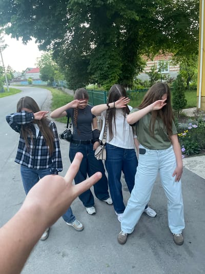 Four young women stand on a paved surface, partially obscured by the hand of the photographer making a peace sign. The women are positioned in a line, facing forward, and each is performing a "dab" pose with their arms covering their faces. The woman on the far left wears a plaid shirt over a white shirt and blue jeans. The second woman from the left wears a grey t-shirt and blue jeans, with a black bag slung over her shoulder. The third woman from the left wears a white t-shirt and blue jeans, with a light-colored bag across her body. The woman on the far right wears a green t-shirt and light blue pants. The background shows a rural setting with trees, a road, houses, and a garden with flowers. The location is identified as Tovstoluh, Ukraine.