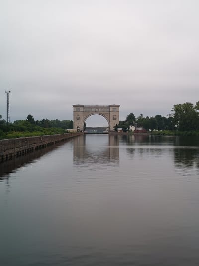 The image depicts a wide canal in Faliukovo, Russia, under an overcast sky. On the left side of the canal, a concrete retaining wall extends into the distance, topped with a metal communication tower. Across the canal, a large, arched structure, likely a hydroelectric dam or a similar water management facility, dominates the scene. This structure is made of stone or concrete and has intricate architectural details. Beyond the arched structure, a dense line of green trees forms the background, with a few small buildings visible near the dam. The water in the canal is calm and reflects the overcast sky and the surrounding environment. The scene appears tranquil and possibly industrial due to the presence of the dam and tower. There are no people or animals visible in the image.