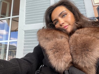 The image is a selfie taken by a woman in a black coat with a large fur collar. The fur is brown, possibly fox. Her long brown hair is loose. She has dark eyebrows and green eyes. She is looking to the right of the camera lens. The image was taken in Roquebrune-Cap-Martin, France, at the coordinates 43.763, 7.463. The background is a wall with white wooden louvers. A window with a view of the city is partially visible on the left side.