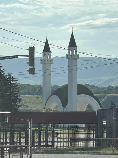 This vertical image captures an outdoor scene in Kubina, Russia, featuring a mosque with two prominent minarets against a backdrop of rolling hills and a sky filled with wispy clouds. The mosque's minarets are white and slender, topped with dark, conical roofs adorned with crescent moons. A large, dark green dome is partially visible between the minarets. In the foreground, to the left, a traffic light is mounted on a pole. To the right, a black metal fence with ornate scrollwork encloses an area, and beyond that, a corrugated metal structure with a small window is visible. The scene appears to be set during the daytime, with bright, diffused light suggesting it could be morning or late afternoon. The weather seems clear, with the sky predominantly blue with white clouds. The overall impression is one of tranquility and architectural beauty in a natural landscape. There is no visible text in the image.