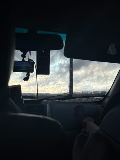 The image, originally oriented sideways, presents a view from inside a vehicle looking out through its windshield. If rotated 90 degrees clockwise to an upright position, the scene reveals a bright, cloudy sky filling the upper part of the windshield, illuminated by strong sunlight, suggesting daytime, likely morning or late afternoon. The clouds exhibit a textured, altocumulus-like pattern.
The vehicle's interior frames this external view. A dashboard or similar panel is visible across the bottom of the windshield. A smartphone or small electronic device is mounted centrally on the windshield or dashboard, connected by wires. Its screen is dimly lit with a blueish hue, but no specific text or images are discernible.
The windshield itself appears dirty or possibly frosted/iced along its lower-right edge and displays a clear crack in the same area. Within the vehicle's interior, in the upper-left corner (if rotated upright), a dark element, possibly a sun visor or part of the ceiling, features a small heart-shaped cutout. In the lower-right section of the interior, a dark car seat or panel is visible, with a light-colored object, possibly a gloved hand, resting upon it. The interior elements are largely in shadow, creating a strong contrast with the brightly lit sky outside. The setting is a vehicle in Aleksandrovskoe, Russia.