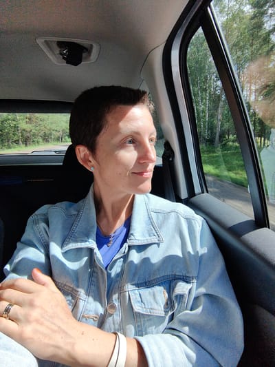 A woman with short, dark hair and blue eyes sits in the passenger seat of a car, looking out the window. She is wearing a light blue denim jacket over a blue t-shirt, and a silver necklace with a small pendant. Her left arm is resting on her lap, and she has rings on two fingers and a white bracelet on her wrist. The car is in motion, as indicated by the blurred trees and greenery visible through the window, suggesting a rural setting. The image appears to have been taken on a sunny day. The location is specified as Svatkaŭski sielśaviet, Belarus.