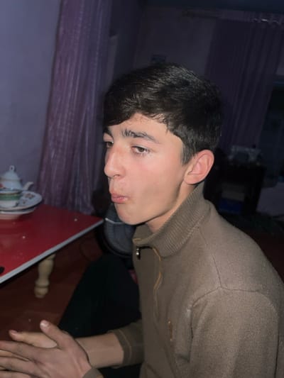 The image presents a close-up, chest-up view of a young male in an indoor setting, captured with dim lighting. The subject's head is tilted downwards, and he is looking towards the lower right of the frame with a pensive or thoughtful expression.

The young man has short, dark brown hair that is styled casually. His skin is fair, and he has distinct dark eyebrows and dark eyes, which are cast downwards. His lips are slightly parted, and a small, light-colored blemish is visible near the left corner of his mouth. He is wearing a long-sleeved, collared sweater or cardigan in a muted brown or beige hue. The garment appears to be made of a knitted material with visible ribbed details on the collar, cuffs, and hem. Two dark buttons or clasps are visible on the front placket of the sweater.

In the background, to the left and extending upwards, a dark purple or maroon fabric, possibly a blanket, duvet, or curtain, with horizontal striped or ridged patterns, is visible. In the lower right corner of the frame, a portion of a red table or flat surface is visible. On this table, a small, white teapot adorned with delicate green and yellow floral patterns is placed next to a matching teacup. A subtle reflection of light can be seen on the glossy surface of the red table. Part of the subject's left hand, with fingers slightly curled, is resting on or near the edge of the table.

The subdued lighting suggests an evening or nighttime setting. The overall mood of the image is quiet and contemplative, with the individual seemingly focused on the tea set or lost in thought. No text is visible in the image.