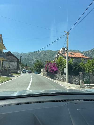 The image depicts a view from a vehicle traveling on a road in Kostanjica, Montenegro. The road curves gently to the right, with a white line marking the center and another white line on the left edge. On the left side of the road, there is a building with a yellow facade and balconies, followed by a rustic stone building with a weathered roof. On the right side of the road, a stone wall borders the property, adorned with vibrant purple bougainvillea. Behind the wall, a stone house with a red tiled roof is visible, along with lush green trees and shrubs. Further in the background, the calm waters of a bay are visible, reflecting the clear blue sky. Majestic, rugged mountains rise in the distance, covered in green vegetation. A few cars are present on the road, including a white van and a dark-colored car. Overhead, utility poles and wires crisscross the sky. The dashboard of the vehicle from which the photo is taken is visible in the foreground. The overall scene suggests a sunny day in a coastal town, characterized by a blend of traditional architecture and natural beauty.
