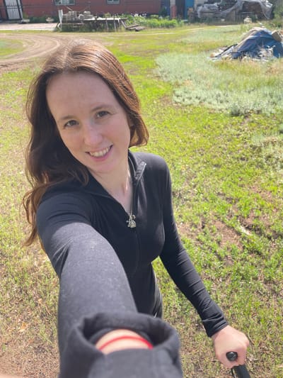 A smiling female, estimated to be in her 20s-30s, with long brown hair and a freckled complexion, takes a selfie outdoors in Vasilevka, Russia. She wears a black long-sleeved top with a partially visible zipper and a silver-colored pendant featuring two small stylized figures on a chain around her neck. A red string bracelet is present on her left wrist, and her right hand holds a black extension pole or similar device, directed towards the camera. The subject is positioned against a bright, sunlit backdrop of a grassy and unpaved area. The ground features a mix of green grass, lighter green low-lying vegetation, and exposed reddish-brown dirt, with a curved dirt path winding through the midground. In the background, a reddish-brick building with windows is visible to the left. To the right, various materials, including stacked wooden pallets and items covered by blue and grey tarps, are accumulated near a smaller structure with blue siding or a blue door. The scene portrays a semi-rural or rural outdoor environment under clear daylight conditions.
