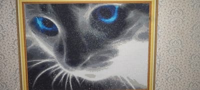 The image shows a framed diamond painting of a cat's face. The cat has striking blue eyes and grey fur. The painting is hanging on a wall with patterned wallpaper. The photograph was taken indoors. There is no information in the image to suggest a location in the city of Svitlovodsk, Ukraine, or any other specific city. The image focuses solely on the artwork and its immediate surroundings.
