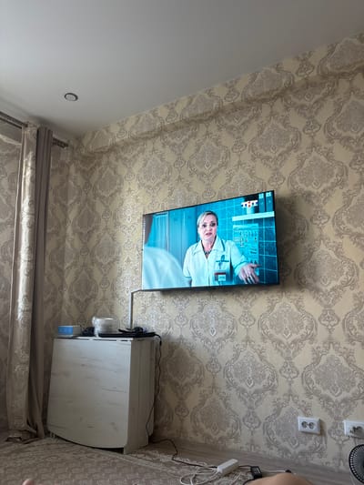 The image shows a wall-mounted television displaying a program. A woman in medical attire is visible on the screen. The wall is covered in patterned wallpaper. To the left of the television, a beige curtain hangs. Below the television, a white folding table is placed against the wall. Various objects, including a laptop, a mouse, and some cables, are on top of the table. The floor is covered with a patterned rug. Electrical outlets are visible on the wall. The scene appears to be an indoor residential setting, possibly a living room, in the city of Nefteyugansk, Russia.