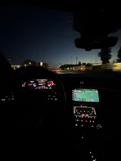 A nighttime view from inside a car traveling on a highway near Melsele, Belgium. The car's navigation system displays a map with a route highlighted in green. The dashboard indicates a speed of 67 km/h. The car's interior is dark, with the rearview mirror visible in the upper left. Traffic lights and other vehicles are visible on the road outside.