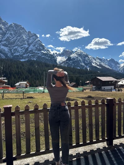 A landscape photograph taken in Sappada, Italy, shows a person with their back to the camera, facing a mountain range. The person is standing behind a wooden fence, wearing a gray long-sleeved shirt and dark jeans, with their hands behind their head. The mountains in the background are partially covered in snow and surrounded by a dense forest. The sky is blue with scattered white clouds. In the midground, there are buildings and structures typical of the Sappada area. The foreground includes a fence and some grassy terrain. The perspective is slightly angled, giving a panoramic view of the scene.
