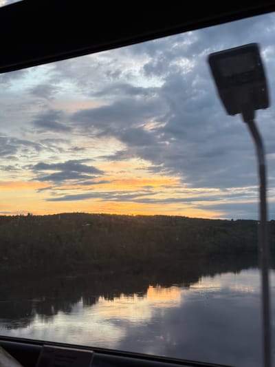 The image shows a view from inside a vehicle, likely a car, looking out towards a body of water reflecting the sky. The sky is filled with clouds, some dark gray and others illuminated by the setting sun, creating a mix of gray, orange, and yellow hues. A treeline is visible on the opposite side of the water. In the foreground, part of a street lamp is visible at the top left, and the frame of the vehicle borders the sides and bottom. The image does not provide information to confirm or deny whether the location is in Arsk, Russia. Therefore, based solely on the visual information, the location cannot be confirmed.