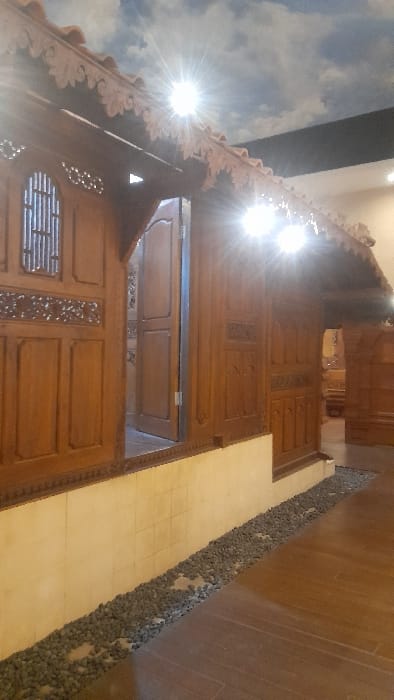 The image displays an indoor scene in Glantengan, Indonesia, featuring a traditional Javanese-style wooden structure. The structure is crafted from dark brown wood, incorporating intricate carvings along its facade and roofline, and rests on a raised base with a light-colored, tiled lower wall. A window with vertical bars and carved detailing is visible on the left. Toward the center, a wooden door is partially open, revealing a glimpse into an interior room. The roof features decorative, carved eaves. Above the structure, the ceiling is a mural depicting a blue sky with white clouds. Several bright, artificial light sources are positioned beneath the roof overhang, producing distinct starburst flares. The ground immediately in front of the structure includes a narrow strip of dark grey pebbles, bordered by the light-colored base wall and a smooth, reddish-brown floor, likely wood, extending to the right. No individuals are present.
