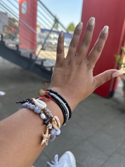 A close-up shot depicts a person's arm and hand adorned with multiple bracelets. The bracelets have various designs, including beads, shells, and a starfish charm. The person's nails are long and light-colored. The background shows urban elements, including metal stairs, a red structure, and partial views of greenery. In the lower right, a portion of a white sneaker is visible. The image was taken in the city of Frechen, Germany.