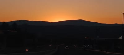 This image depicts a sunset over a mountainous landscape, likely near La Chopera, Spain. The sky is a vibrant gradient of orange and yellow, with the sun setting behind a dark silhouette of mountains in the distance. In the foreground, there is a dimly lit highway with several cars driving. On the right side of the image, a Repsol gas station is visible, also silhouetted against the fading light. The overall scene is one of twilight, with the natural beauty of the sunset juxtaposed with elements of human infrastructure.