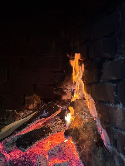 A close-up shot of a fire burning inside a brick fireplace. Flames are visible between the logs, and embers glow within the wood. The bricks of the fireplace appear dark. The location in the city of Lilkovo, Bulgaria, is not discernible from the image.