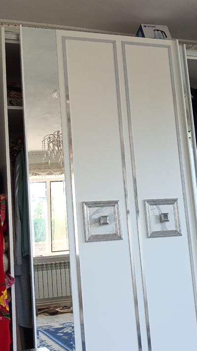 The image shows a white wardrobe with mirrored doors and decorative silver trim. There are two square decorative elements with a smaller square in the center on the right-hand doors. To the left of the wardrobe, a closet is partially visible, revealing clothing and a box with a "VITEK" logo on top of the wardrobe. A window with a radiator below it is visible in the mirror reflection. The floor has a blue and white patterned rug. The overall scene suggests an indoor domestic setting. It is difficult to ascertain the exact location in Maqat, Kazakhstan based on this image alone.