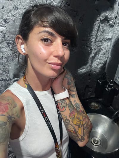 An adult female is depicted in a selfie, positioned indoors in the city of Benoa, Indonesia. She possesses dark hair styled with bangs, brown eyes, and is looking directly at the camera with a slight smile. A white wireless earbud is present in her left ear. She wears a white ribbed tank top and a black lanyard around her neck, branded "VAXXO" with accompanying text "IMAGINED ART EXPERIENCE" and a colorful graphic. Extensive tattoos featuring floral patterns are visible on her left arm, and multiple colorful, cartoon-style designs are present on her right arm. A white smartwatch is worn on her right wrist, with her right hand gently touching her neck/jawline. To her right, on a dark countertop above a silver metal basin, stand two black pump bottles labeled "CREAM" and "SOAP." The background features a dark, roughly textured wall with gray and silver highlights.