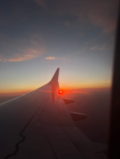 The image shows an aerial view from an airplane window during sunset. The airplane wing occupies the foreground, with the Ryanair logo visible on its tip. The sun is a bright, red orb just above the horizon, casting a warm glow on the clouds and the wing. The sky transitions from a deep blue at the top to shades of orange and pink near the horizon. Faint contrails from other aircraft are visible in the sky. The scene suggests an aircraft is in flight, likely over a landscape illuminated by the setting sun. The location in Póvoa de Lanhoso, Portugal, is inferred, as this is a popular flight route.