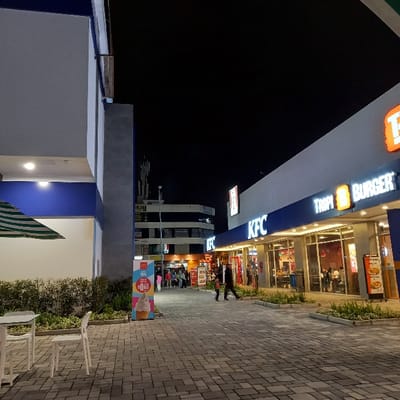 This nighttime image depicts a commercial area in Tumbaco, Ecuador, illuminated by artificial light. On the right, a single-story building houses two fast-food establishments: KFC and Tropi Burger, both featuring prominent, illuminated signage and glass storefronts revealing customers inside. A paved walkway of gray interlocking bricks occupies the foreground and midground, leading towards additional commercial structures in the distance. To the left, a portion of another building with a blue and gray facade is visible, alongside an outdoor seating area featuring a white table, two white chairs, and a partially open green and white striped umbrella. Low-lying green landscaping borders the buildings along the walkway. Approximately 10-15 individuals are visible; two adults walk past KFC towards the camera, while others are seen inside the restaurants or congregating further down the walkway near additional businesses. A freestanding McDonald's advertisement for ice cream, priced at $0.65, is positioned on the left side of the walkway. In the background, beyond the immediate commercial strip, a multi-story building with multiple antennas or communication towers on its roof is discernible against the dark sky.