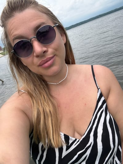 A woman is taking a selfie outdoors by a body of water. She has blonde hair and is wearing sunglasses and a black and white striped dress. She is also wearing a white beaded necklace. The water appears to be calm, and the sky is cloudy. There is a treeline visible in the distance across the water. The image does not provide enough information to determine the specific location within Nikol'skoe, Russia.