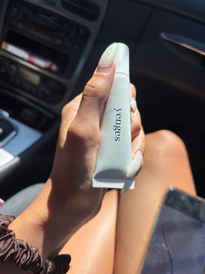 An individual's hand, adorned with glittery, almond-shaped nails, holds a small, white tube of product with the word "yenges" printed vertically in black text. The person is seated in the passenger seat of a car, as indicated by the dashboard and car interior visible in the background, possibly on a sunny day in Binkos, Bulgaria. Sunlight streams into the car, casting shadows. A brown scrunchie is visible on the person's wrist. A smartphone lies on the person's lap, its screen displaying a blue-tinted image, possibly a map.