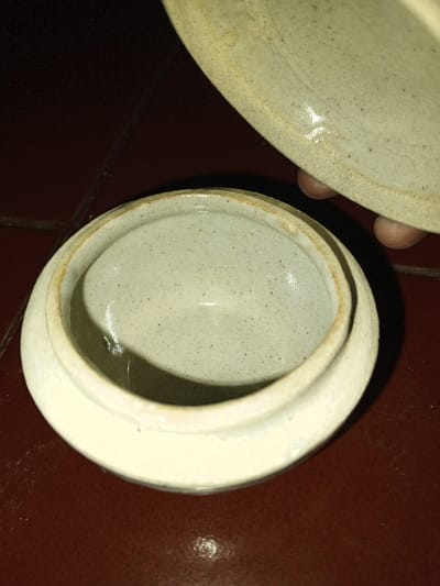 An overhead view captures a speckled light gray ceramic jar, open, resting on a dark reddish-brown surface, possibly a tiled floor, in Longmiang, Laos. The round jar features a slightly flared rim showing minor discoloration. Its interior is visible and empty. A human hand, with fingers partially visible on the right, holds the matching speckled light gray ceramic lid elevated above the jar, indicating the act of opening or closing. The scene is dimly lit.