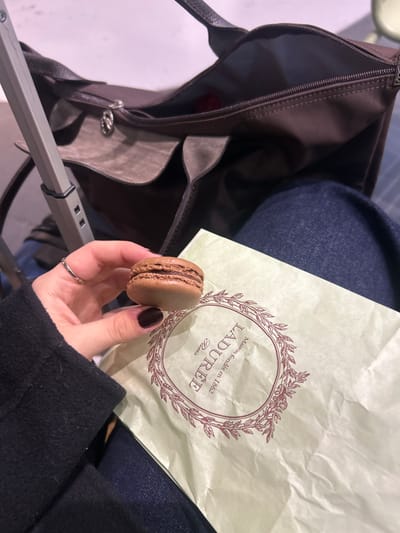 An individual is seated, likely within a waiting area of Charles de Gaulle Airport (CDG) in Tremblay-en-France, France. A left hand, adorned with dark nail polish and a silver ring on the ring finger, holds a single brown macaron with a visible chocolate-colored filling. A crumpled light green Ladurée paper bag, featuring the brand's logo and "Paris Maison fondée en 1862," rests on the person's lap, which is clad in dark blue denim jeans. Adjacent to the lap is an open, dark brown handbag or travel bag, containing an indistinct red object. A grey telescopic handle of a piece of rolling luggage is partially visible on the left side, positioned upright next to the handbag. The background is a soft, indistinct light tone, consistent with an indoor public space.
