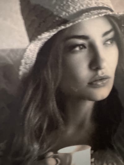 A black and white, low-light image depicts a close-up of a person, likely a young adult female, oriented as if looking upwards. Her face is seen in profile from the nose up, with her eyes directed towards the top of the frame, conveying a pensive or contemplative expression. She has long, dark hair visible towards the bottom right and appears to be wearing a textured, possibly knitted or woven, hat or hood that frames the left side of her face. Her lips are slightly parted.

On the right side of the image, a small, light-colored cup or container is visible, held by her hand, suggesting she is about to drink or is holding a beverage. The background is dark and largely out of focus, creating a sense of depth and emphasizing the subject. The soft focus and grainy texture contribute to an intimate and quiet mood, indicative of subdued lighting, possibly indoors or during the evening. There is no visible text or distinct location cues within the image itself. The provided context indicates the location as Troisvierges, Luxembourg.