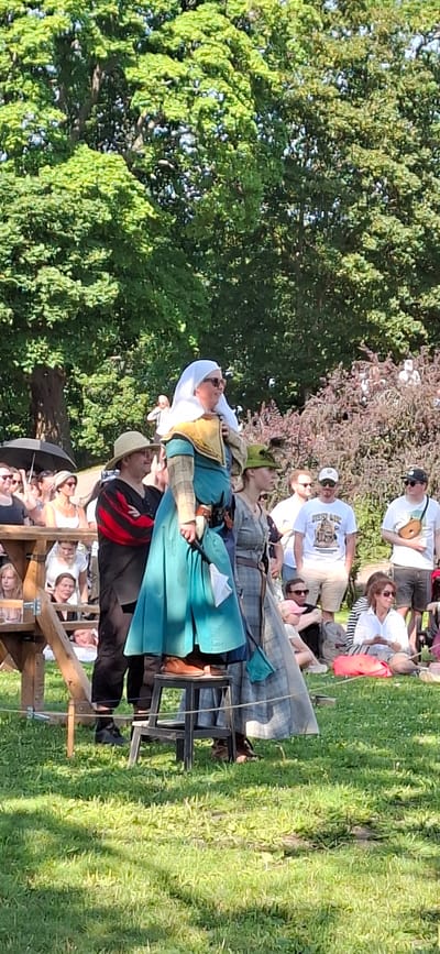 A full outdoor shot captures a sunny day, likely in Turku, Finland, given the context of a historical reenactment. In the foreground, a person dressed in historical garb, featuring a turquoise gown and a white head covering, stands elevated on a stool. They are looking to the right, with their hand on their chest. To their left, another individual in a wide-brimmed hat and a black garment with red stripes stands beside them. Behind these central figures, a crowd of spectators are visible, some seated on the grass and others standing. The environment includes lush green trees in the background and a wooden structure on the left side of the frame. The overall atmosphere suggests a public event or festival.