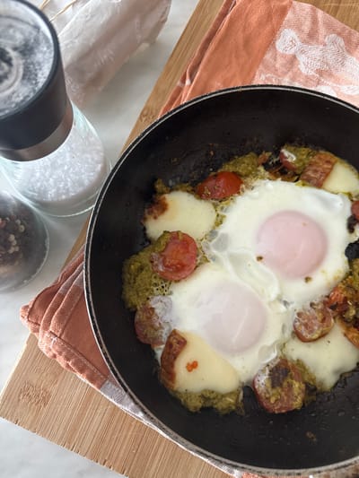 This image depicts a close-up, overhead view of a black frying pan containing a cooked meal, situated on a light-colored kitchen countertop in the Kalyvia Thorikou Municipal Unit, Greece. The pan holds two fried eggs with fully set whites and pale pinkish yolks. Surrounding the eggs is a textured green pesto-like sauce, interspersed with halved red cherry tomatoes and several irregular pieces of melted white cheese, some exhibiting slight browning.
The frying pan rests on a light brown wooden cutting board, which is partially covered by a folded orange and white striped or checkered kitchen towel on the left side. To the left of the pan, two glass grinders are visible: one is transparent and filled with coarse white salt, and another, partially obscured, appears to contain dark peppercorns. A crumpled piece of light-colored paper or fabric is visible in the upper background. There are no people, actions, or interactions depicted. The scene is brightly lit.
