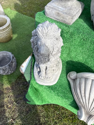 The image depicts a collection of stone garden ornaments displayed on artificial green turf. Dominating the scene is a lion statue, viewed from above, its head and paws clearly visible. Adjacent to the lion are other stone elements, including architectural fragments and a vase or planter. The setting appears to be outdoors, with natural grass and soil visible around the artificial turf, suggesting a garden or display area. The image's origin in Chirk, United Kingdom, implies this could be a garden center or a residential garden in that locale.