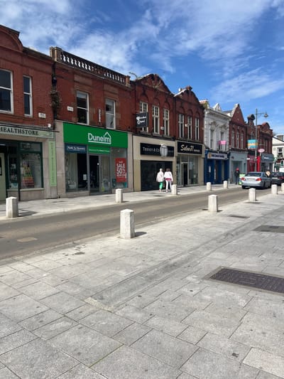 The image shows a street scene in Dún Laoghaire, Ireland, with shops lining one side of the street. The architecture appears to be traditional, with red brick buildings and white trim. Several storefronts are visible, including Dunelm, Tierney & Co, Salon 71, and the Irish Cancer Society. A silver sedan is driving on the road, and a person is standing on the sidewalk. The sky is blue with some white clouds. The street is paved with cobblestones, and there are several stone bollards separating the sidewalk from the road. The overall impression is a typical street scene in a commercial district.
