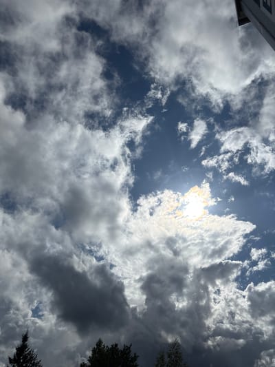 A low-angle, full shot captures a dramatic sky filled with a mix of dark, heavy clouds and brighter, wispy formations. The sun peeks through, creating a bright, slightly overexposed area with rainbow-like iridescence. In the foreground, silhouetted evergreen trees are visible at the bottom edge of the frame, suggesting an outdoor, possibly wooded, location. A portion of a building's facade, featuring white and grey panels, is visible in the upper right corner. The scene appears to be captured in Sauleskalns, Latvia, under a dynamic sky.