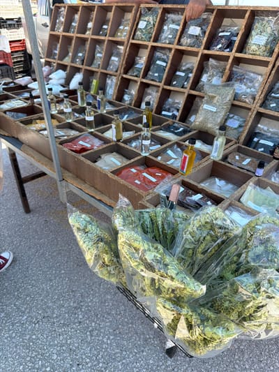 In Ασπροβάλτα, Greece, a vendor displays a market stall filled with various herbs and spices. The stall features a wooden rack with numerous compartments, each containing small plastic bags of dried herbs and spices, some in bulk and others in more refined forms. Several small glass bottles, likely containing oils or tinctures, are also visible on the stall. In the foreground, large, clear plastic bags are filled with dried green herbs, possibly oregano or thyme, presented in a basket. The ground is paved with asphalt, and a portion of a person's leg wearing a red sneaker is visible on the left side of the frame. In the background, crates and a portion of a vehicle can be seen, suggesting an outdoor market setting. The overall scene depicts a typical Mediterranean herb and spice market.