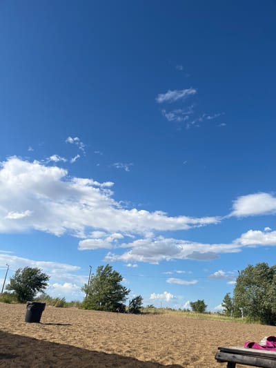 A wide shot captures a sandy area with sparse vegetation under a bright blue sky dotted with white clouds. In the foreground, a black trash can stands to the left, casting a long shadow. A wooden bench is partially visible in the lower right corner, with what appear to be a pair of pink slippers on it. In the middle ground, several trees with green foliage are scattered across the landscape, and a few individuals are seated or lying down on the sand in the distance to the right. Poles with lights are visible in the background. The scene suggests an outdoor recreational area, possibly a beach or park, in Gres-2, Kazakhstan.