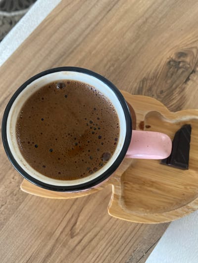 A close-up, overhead shot shows a mug of Turkish coffee with a frothy top, placed on a wooden saucer. A piece of dark chocolate rests on the saucer next to the mug. The mug has a pink handle and a black rim. The wooden saucer has a light-colored grain and is shaped with a notched edge. The coffee and saucer are on a wooden table with a distinct grain pattern. The image appears to be taken indoors.