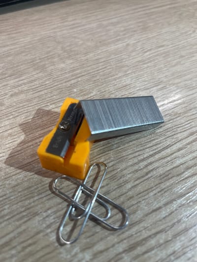 The image displays a close-up, eye-level view of office supplies on a wooden surface. The primary subjects are a yellow pencil sharpener with a metal blade, a stack of silver staples, and two silver paper clips. The pencil sharpener has the brand name "Erich Kraus" inscribed on its blade. The wooden surface appears to be a table or desk, with a visible wood grain pattern. No people or animals are present in the image, and no specific city location is identifiable from the visual information. The lighting suggests an indoor setting, possibly with artificial light.